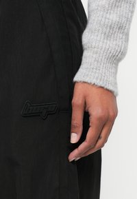 Pantaloni negri dintr-un material texturat, având o inscripție logo neagră bine definită. O mână atinge blând cusătura laterală, evidențiind detaliile materialului.