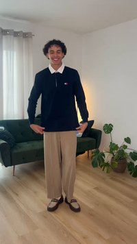 Νεαρός άνδρας που χαμογελά, φοράει μαύρο πουκάμισο με μακριά μανίκια και μπεζ φαρδιά παντελόνια, στέκεται σε φωτεινό σαλόνι με πράσινο καναπέ και φυτά.