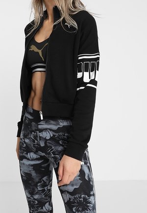 Frau trägt schwarze Puma-Zip-Jacke, schwarzen Sport-BH mit goldenem Puma-Logo und schwarz-grau gemusterte Leggings mit Blumenmuster.