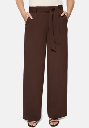 Pantalon marron taille haute à jambe large avec ceinture en tissu nouée à la taille, porté avec des sandales noires à lanières et les mains dans les poches.