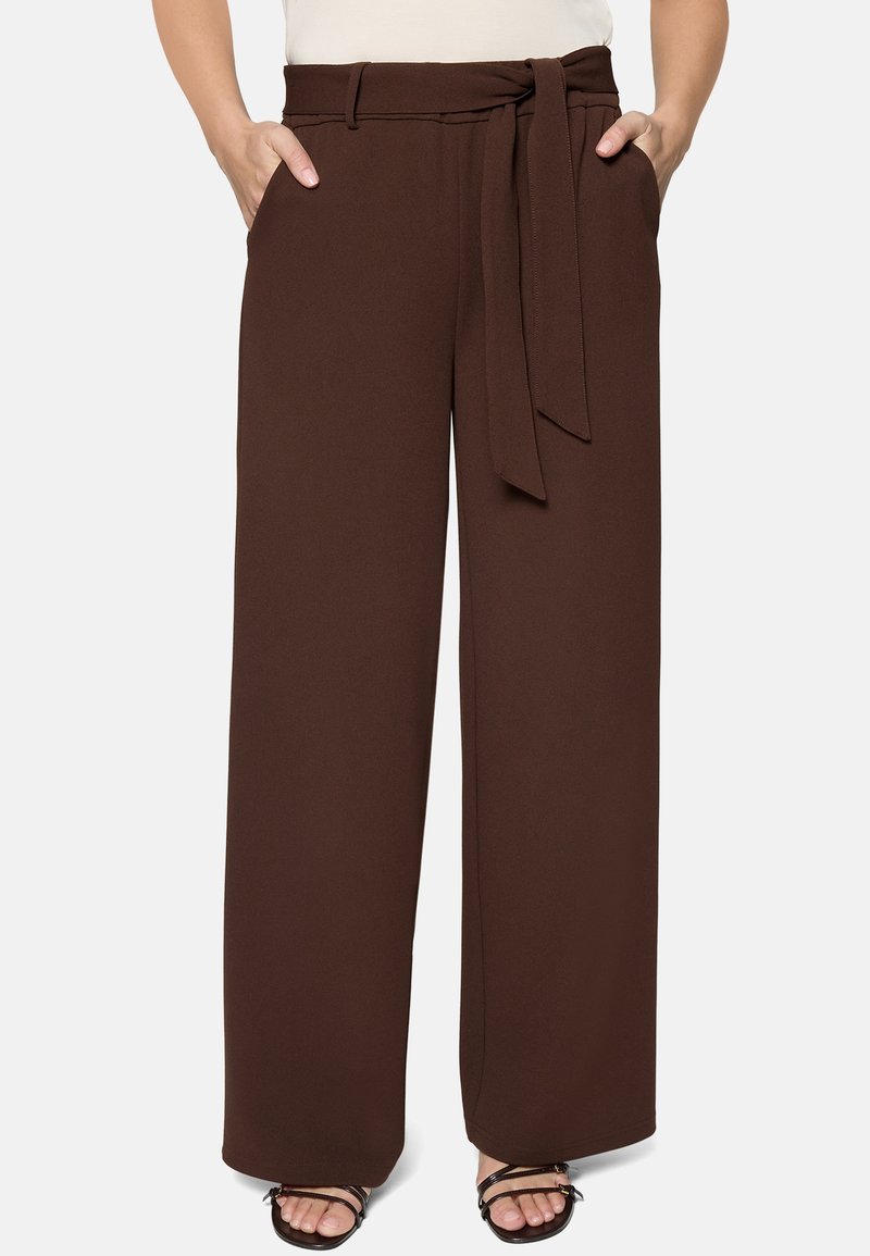 Pantalon marron taille haute à jambe large avec ceinture en tissu nouée à la taille, porté avec des sandales noires à lanières et les mains dans les poches.