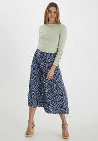 Hellgrünes, geripptes Langarmoberteil mit drei Knopfakzenten, kombiniert mit einem blauen, blumigen Midikleid und beigen Keilsandalen.