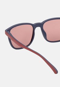 Gafas de sol con montura mate en azul oscuro y lentes gradientes en tono rosa. Acento rojo en la delgada patilla con el logotipo "GANT" grabado.