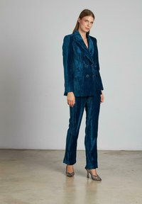 Abito doppiopetto turchese in un tessuto lucido e strutturato; presenta pantaloni abbinati e scarpe con tacco alto metallizzate per un look coordinato.