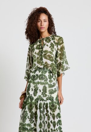 Femme aux cheveux bouclés portant une robe blanche avec des motifs feuillus verts ornés et des manches transparentes, debout devant un fond clair uni.