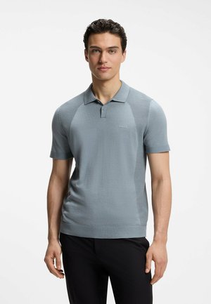 Junger Mann, der ein hellblaues kurzärmeliges Polohemd mit dem "BOSS"-Logo trägt, steht mit entspannten Händen vor einem schlichten weißen Hintergrund.