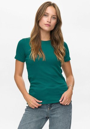 Frau mit langem, welligem Haar, die ein tailliertes, kurzärmliges T-Shirt in Türkis und eine blaue Jeans trägt, vor einem schlichten weißen Hintergrund stehend.