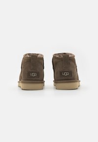 Bottines en suède marron avec doublure douce, bout arrondi et semelle en caoutchouc basse. Présente un logo UGG sur le talon et une boucle à l'arrière pour un enfilage facile.