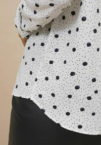 Weiße Bluse mit einem Muster aus marineblauen Punkten und kleineren schwarzen Punkten. Die Bluse hat einen abgerundeten Saum und voluminöse Ärmel.