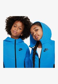 Kaksi lasta wearing matching sinisissä Nike-huppareissa, joissa on mustat vetoketjut ja logot, sileä kangas ja huput.