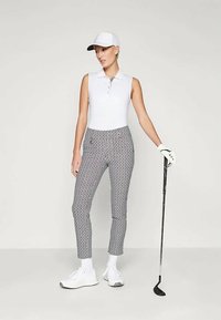 Balta bez piedurknēm polo krekls, rakstaini pelēki golfa bikses, balta cepure, baltas cimdi un sporta apavi. Modelis tur golfa nūju, minimālistisks fons.