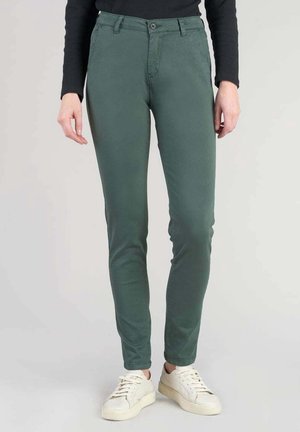 Persona que lleva pantalones ajustados verde oscuro, camiseta de manga larga negra y zapatillas blancas, de pie contra un fondo liso.