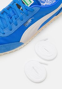 Kék sportcipő velúr és textil anyagból, fehér oldalsávval, textúrált talppal, mellette egy kiegészítő fehér fűzővel.