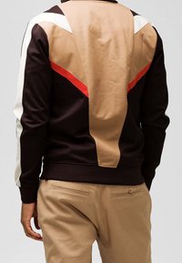 Tummanruskea collegepaita, jossa on beige, valkoinen ja punainen väri-blokattu muotoilu selässä. Paita on sileäpintainen ja siinä on ribbitoitu helma. Yhdistetty vaaleisiin housuihin.