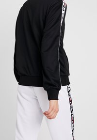 Veste de sport noire avec bordure blanche et détail de logo sur les manches, associée à un pantalon blanc avec une bande verticale à logo. Texture de tissu lisse.