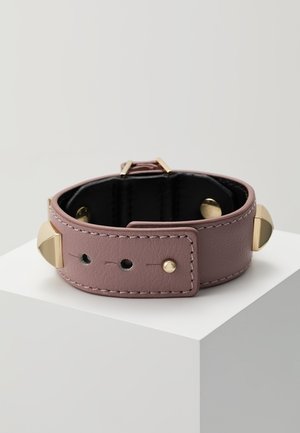 Bracelet en cuir mauve discret avec des clous pyramidaux dorés et une boucle, exposé sur un socle blanc sur fond clair.