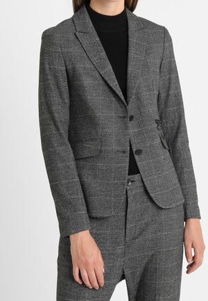 Mujer que lleva un blazer gris a medida de cuadros y pantalones a juego sobre un jersey de cuello alto negro.
