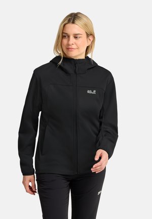 Donna con giacca nera con cappuccio e pantaloni neri, in piedi davanti a uno sfondo chiaro e uniforme, che guarda leggermente di lato.