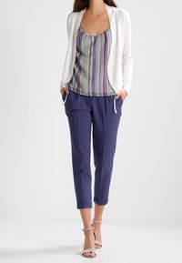 Cárdigan blanco abierto por delante sobre una camiseta sin mangas de rayas multicolores, combinado con pantalones tapered azul marino y tacones gris con tira en el tobillo.