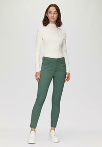 Weißes, tailliertes Langarmoberteil mit vertikalen Nähten, kombiniert mit grünen Slim-Fit-Hosen. Das Model trägt weiße chunky Sneakers und steht vor einem neutralen Hintergrund.