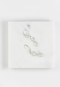 Orecchini argento a forma di infinito con pietre gemme trasparenti, sospesi a chiusura a leva, esposti su una superficie in marmo.