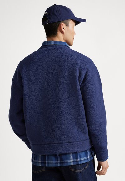 Uomo con giacca in pile blu navy, camicia a quadri sotto e cappellino blu navy, visto di spalle su sfondo bianco uniforme.