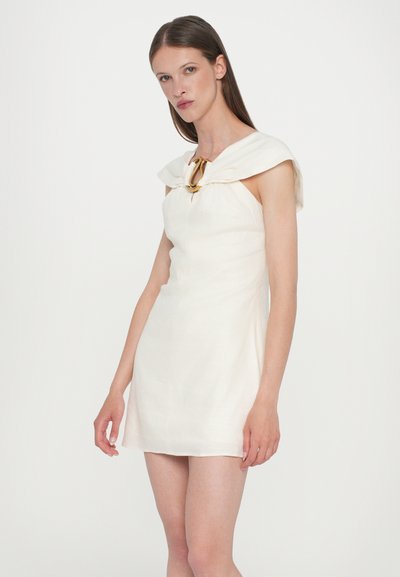 Short cream dress with a structured, draped neckline, cinched bodice, and a gold accent at the collar. Smooth texture, tailored fit.