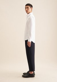 Camicia bianca a maniche lunghe, pantaloni blu scuro e scarpe nere. Polsini abbottonati e colletto strutturato. Tessuto liscio con una leggera lucentezza.