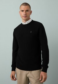 Hombre vestido con un suéter negro texturizado sobre una camisa blanca de cuello y pantalones beige, de pie contra un fondo gris claro.