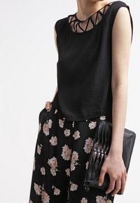 Top negro sin mangas con un intrincado escote recortado, combinado con pantalones negros con estampado floral. Cartera de mano negra con pliegues.