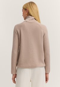 Helles rosa geripptes Strickoberteil mit hohem Kragen, langen Ärmeln und lockerer Passform, das eine strukturierte Oberfläche aufweist.