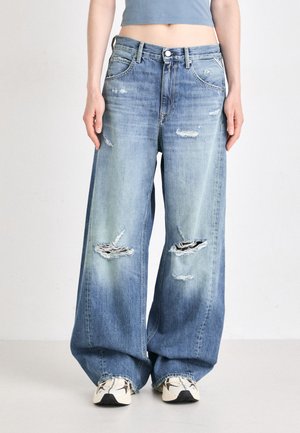 Jeans ampi di denim azzurro chiaro con toppe strappate, dotati di un'apertura con bottoni, tasche e una vestibilità ampia. Abbinate a scarpe da ginnastica beige.