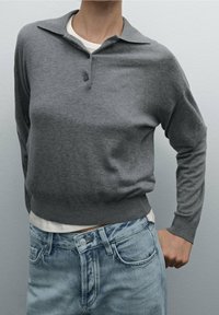 Harmaa neulepusero, jossa on kaulus, kahden napin suljenta ja pitkät hihat. Yhdistetty vaaleansinisiin farkkuihin.