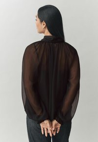 Čierna priehľadná blúzka s dlhými, mierne naberanými rukávmi, nariasením pri golieri a elastickými detailmi na zápästiach. Nosená s tmavými džínsy.