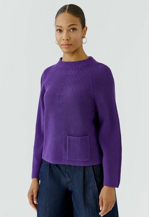 Vrouw die een paarse gebreide trui met een voorzak en donkerblauwe spijkerbroek draagt, staand tegen een effen lichte achtergrond.