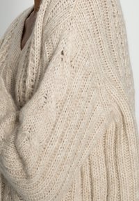 Pull en tricot beige clair avec col en V, motif torsadé et texture côtelée. Caractérisé par des manches larges et amples ainsi qu'une apparence confortable.