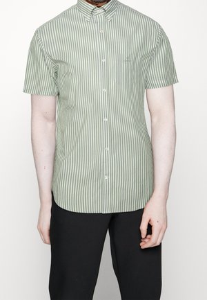 Hombre que lleva una camisa de manga corta con rayas verticales verdes y blancas, abotonada y con un bolsillo en el pecho, y pantalones negros.