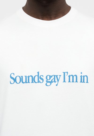 Άνδρας με άσπρο μπλουζάκι και μπλε κείμενο στο στήθος που λέει «Sounds gay I'm in», εστιασμένος στο πάνω μέρος του κορμού και στη γραμμή του λαιμού.