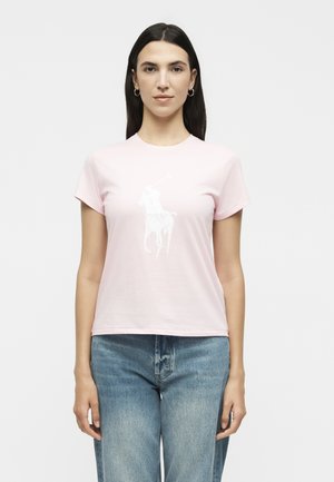 Γυναίκα με μακριά σκούρα μαλλιά φοράει ροζ ανοιχτό T-shirt με λευκό λογότυπο παίκτη πόλο και μπλε τζιν, στέκεται μπροστά σε απλό λευκό φόντο.