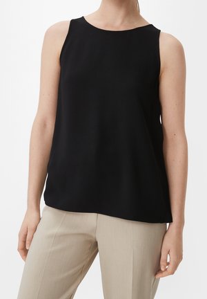 Haut noir sans manches porté avec un pantalon beige taillé sur mesure sur une personne debout devant un fond blanc.