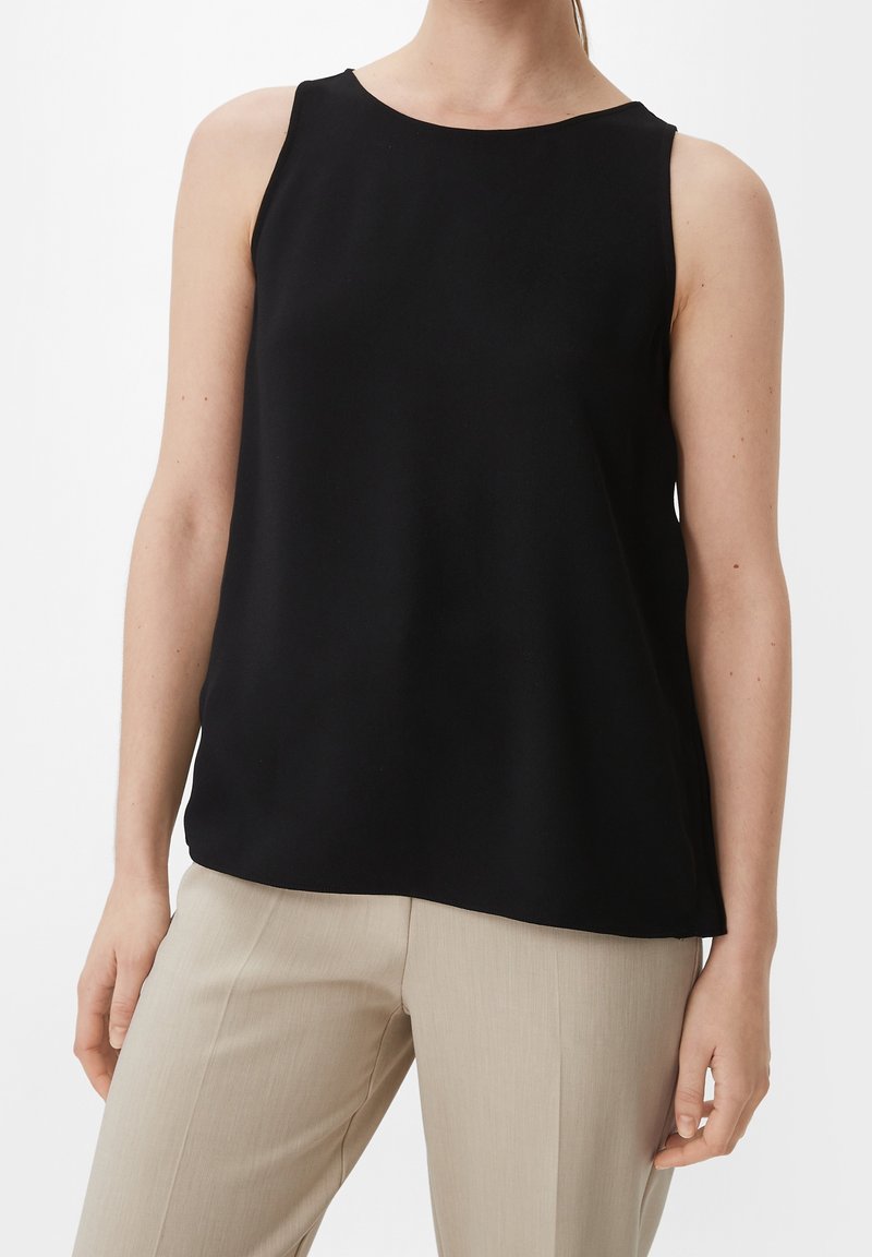 Haut noir sans manches porté avec un pantalon beige taillé sur mesure sur une personne debout devant un fond blanc.