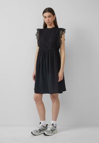 Robe noire avec un haut en dentelle et des manches volantées, taille cintrée et jupe fluide. Assortie avec des baskets argentées et des chaussettes blanches.