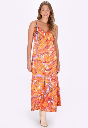 Fiatal nő mosolyog, hosszú, ujjatlan paisley mintás ruhát visel élénk narancssárga, lila és rózsaszín árnyalatokban, bézs bokacsizmával és narancssárga fülbevalóval.