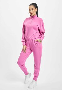 Felpa rosa con colletto alto e zip a metà, abbinata a pantaloni jogger coordinati, entrambi realizzati in un tessuto morbido; completata con sneakers bianche.