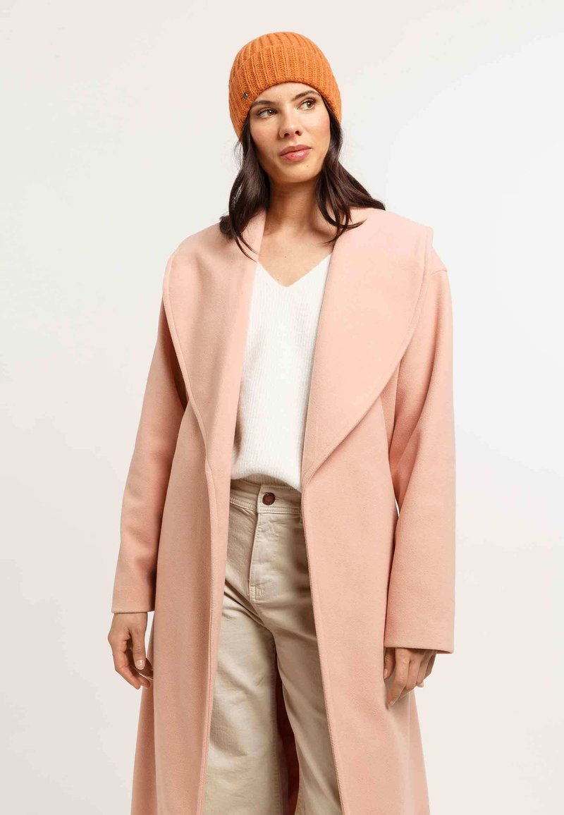 Palton roz din lână lungă cu rever larg, purtat cu un top alb din tricot și pantaloni bej. O căciulă din tricot cu nervuri portocalie oferă un contrast.