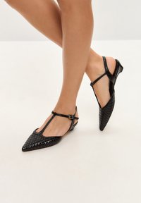 Pantofi din piele țesută negri, cu vârf ascuțit, curele subțiri la glezne și toc jos, având o textură netedă și un design minimalistic.