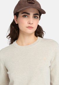 Maglione beige chiaro con scollo rotondo, morbida texture e un piccolo logo beige. Cappello in corduroy marrone scuro con logo ricamato.