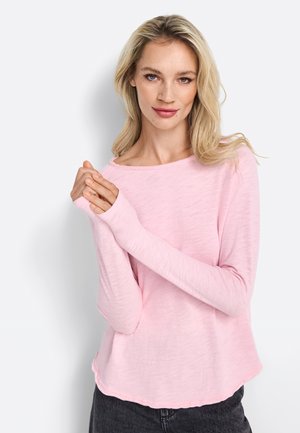 Helles rosa Langarmshirt mit Bootsausschnitt und leicht strukturierter Stoff. Der Saum hat eine subtile wellenförmige Kante. Die Trägerin hat die Hände gefaltet.