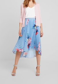 Cardigan rosa chiaro su una maglietta bianca a scollo a V, abbinato a una gonna plissettata blu con grandi fantasie floreali in rosa e viola. Tacchi argentati.