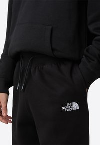 Svart hettejakke med frontlomme og svarte joggebukser. Har elastisk midjebånd med snorer og hvit brodert logo på buksen.